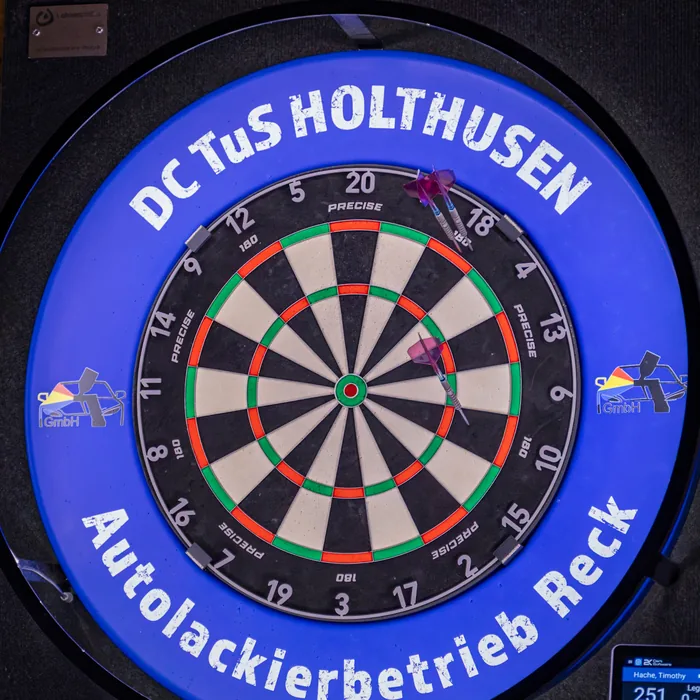 Dartboard mit dem Schriftzug DC TuS Holthusen, drei Pfeile stecken im Board, rechts das Anzeigegerät