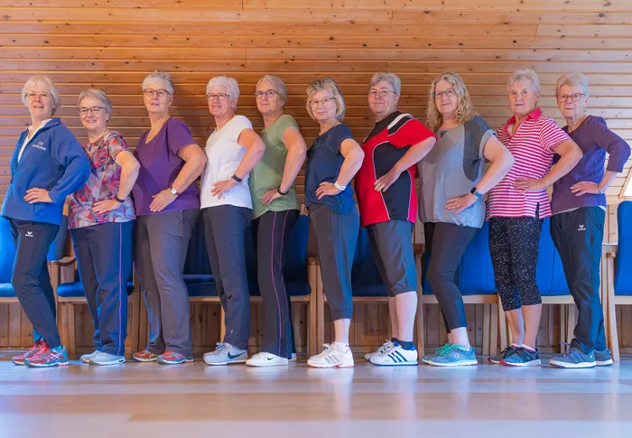 Zehn Frauen der Hausfrauen-Gymnastik in einer Reihe im Vereinsheim, hinter ihnen die Holzwand und die blauen Stühle des Saals